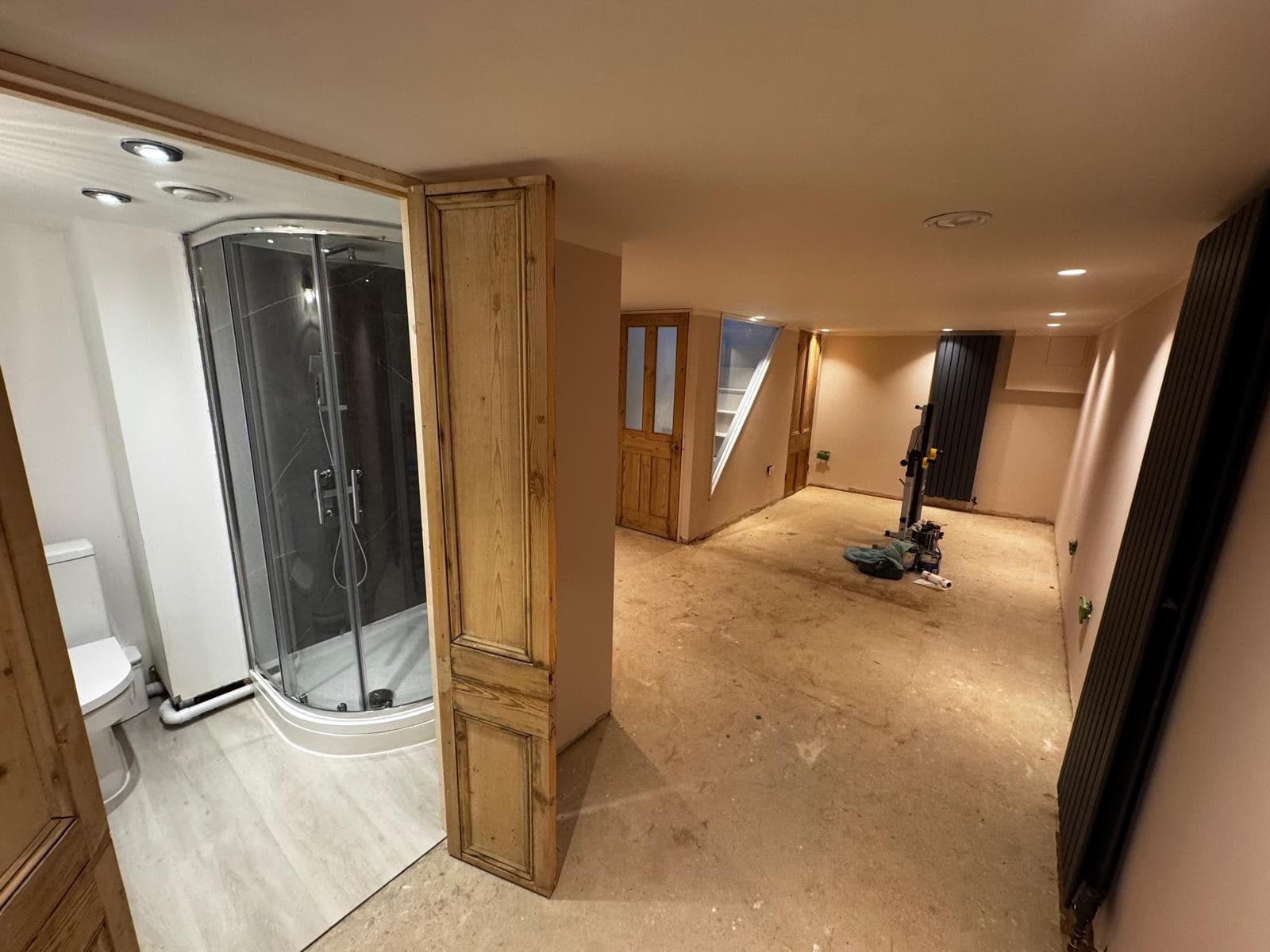 Finished shower room with a glazed quadrant enclosure, lit and decorated, seen from the main basement room before flooring