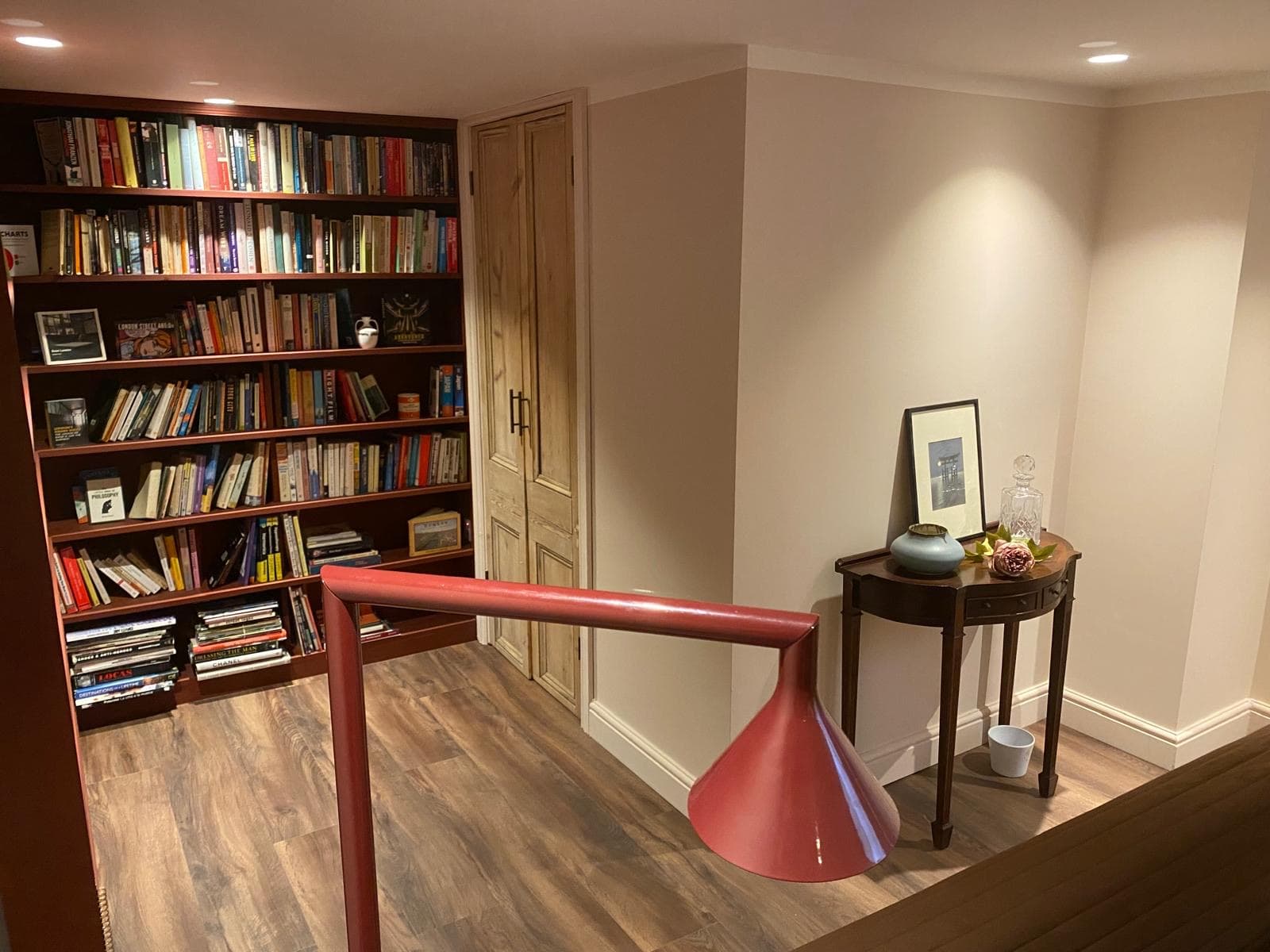 Finished basement library with a full height bookcase, red handrail and a console table under warm downlights